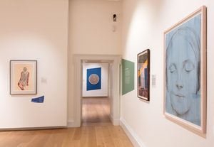 An interior view of a museum gallery with several framed artworks hung on the walls, including figurative drawings, a circular abstract piece, and a large portrait of a face.