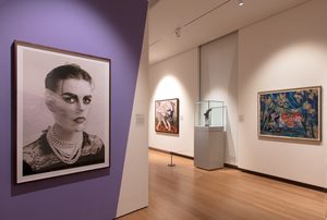 A gallery space exhibiting framed artworks including a large portrait, paintings, and a sculptural piece encased in glass. The walls are purple-coloured.