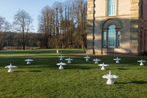 Multiple white mushroom-shaped sculptures are arranged on a grassy area leading up to a large stone mansion with bare trees in the background.
