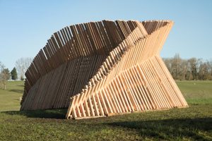A massive curved wooden structure made of parallel slats stands in an open grassy field against a clear blue sky.