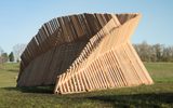A massive curved wooden structure made of parallel slats stands in an open grassy field against a clear blue sky.