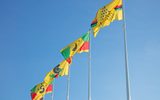 Five tall flagpoles fly bright yellow flags emblazoned with a design of red, green, and black stripes, stars, and triangular patterns against a clear blue sky.
