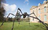 A large, spider-like metal sculpture with outstretched spindly legs stands on the lawn in front of an ornate stone building with arched windows.