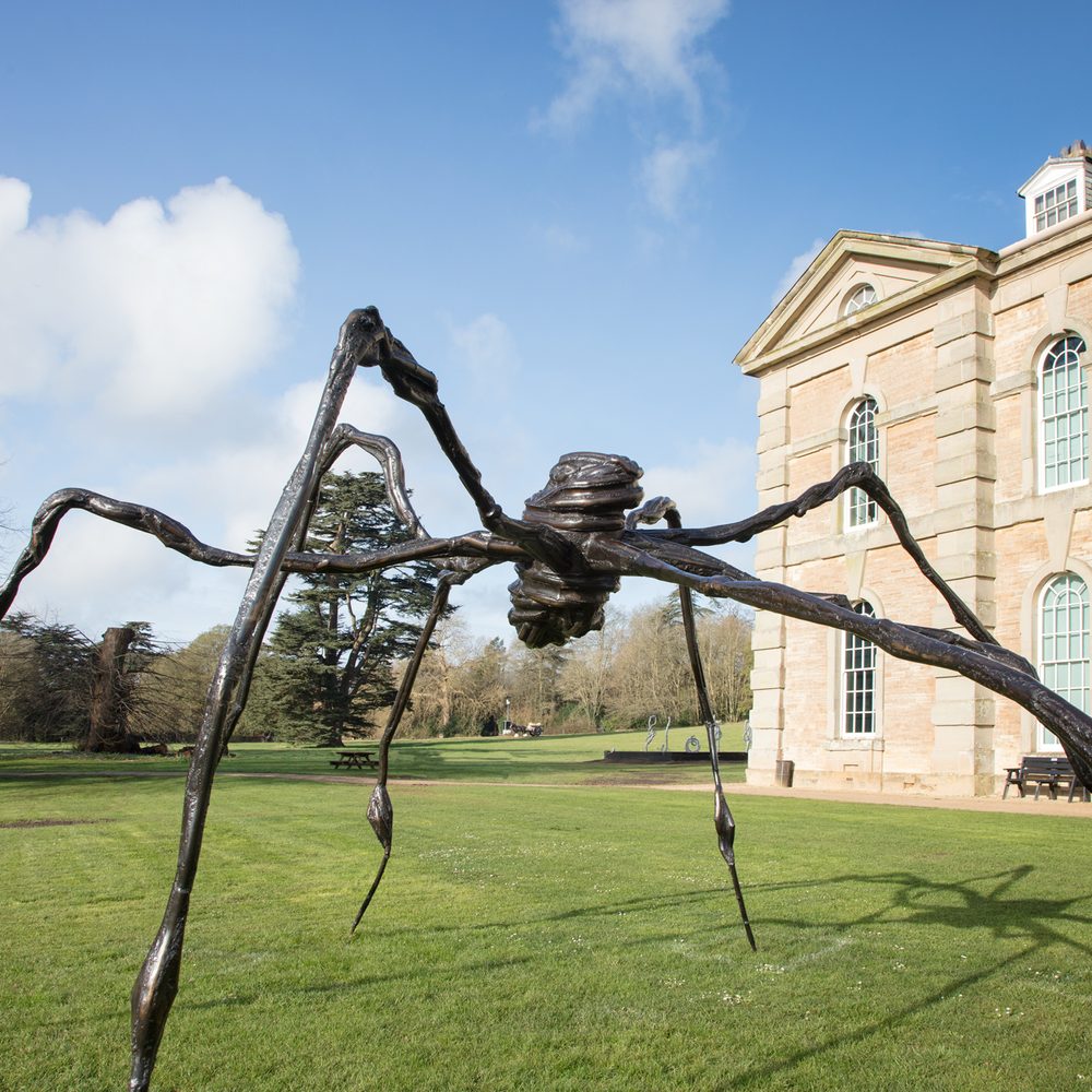 A large spider sculpture in a green meadow below a blue sky next to a neoclassical building.