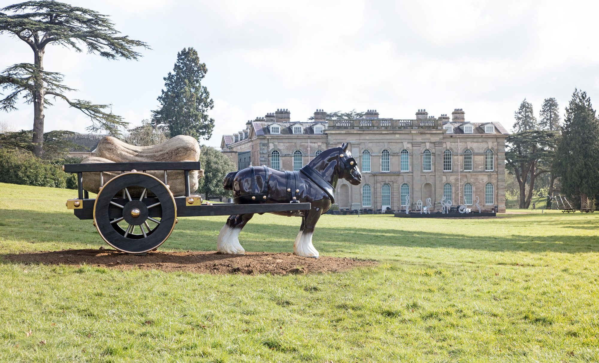 A sculpture of a horse and carriage stands in a meadow overlooking a neoclassical building
