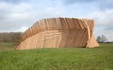 A massive curved wooden structure made of parallel slats stands in an open grassy field against a cloudy sky.