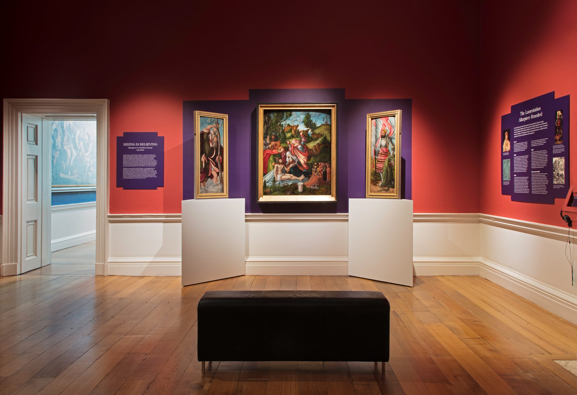 An art gallery room with red and purple walls, displaying several framed paintings and exhibits. The central painting appears to depict a biblical or mythological scene with figures in a lush, forested setting. A bench is placed in the centre of the room for viewing the artworks.