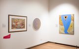 A section of a museum gallery with framed artworks hung on the walls. The works include a colourful abstract painting, a circular sculpture, and a Renaissance-style figurative painting.