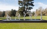 A series of abstract silver fountain sculptures resembling intestines in a shallow pool stand in a row in a grassy park setting, with tall trees in the background.