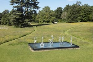 0U9A4878-Edit An outdoor water feature with abstract white sculptural forms emerging from a square reflecting pool, surrounded by grass and wildflowers.
