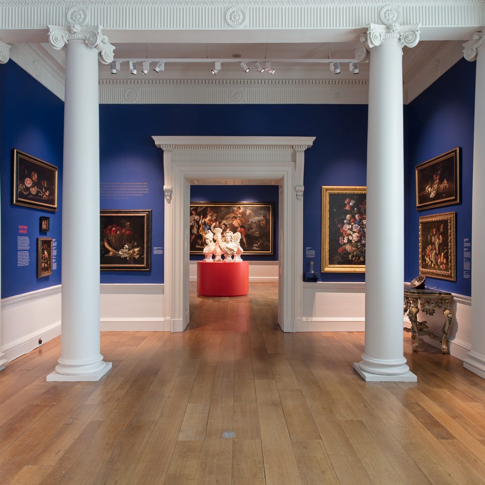 Looking towards a door and two white collums in a blue art gallery with Neapolitan paintings and sculpture on the walls
