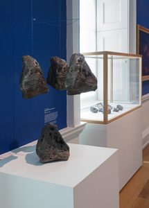 This image shows an art installation in a blue art gallery setting. Three bronze casts of magma rock hang above the original piece. Next to them is a glass display case filled with similar rocks.
