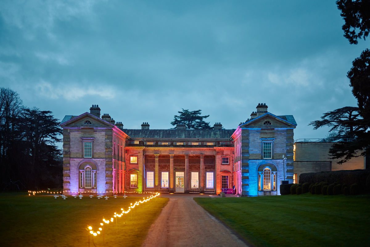 Compton Verney, a neo-classical house with columns in a green meadow and long drive, is lit up at dusk with many colours