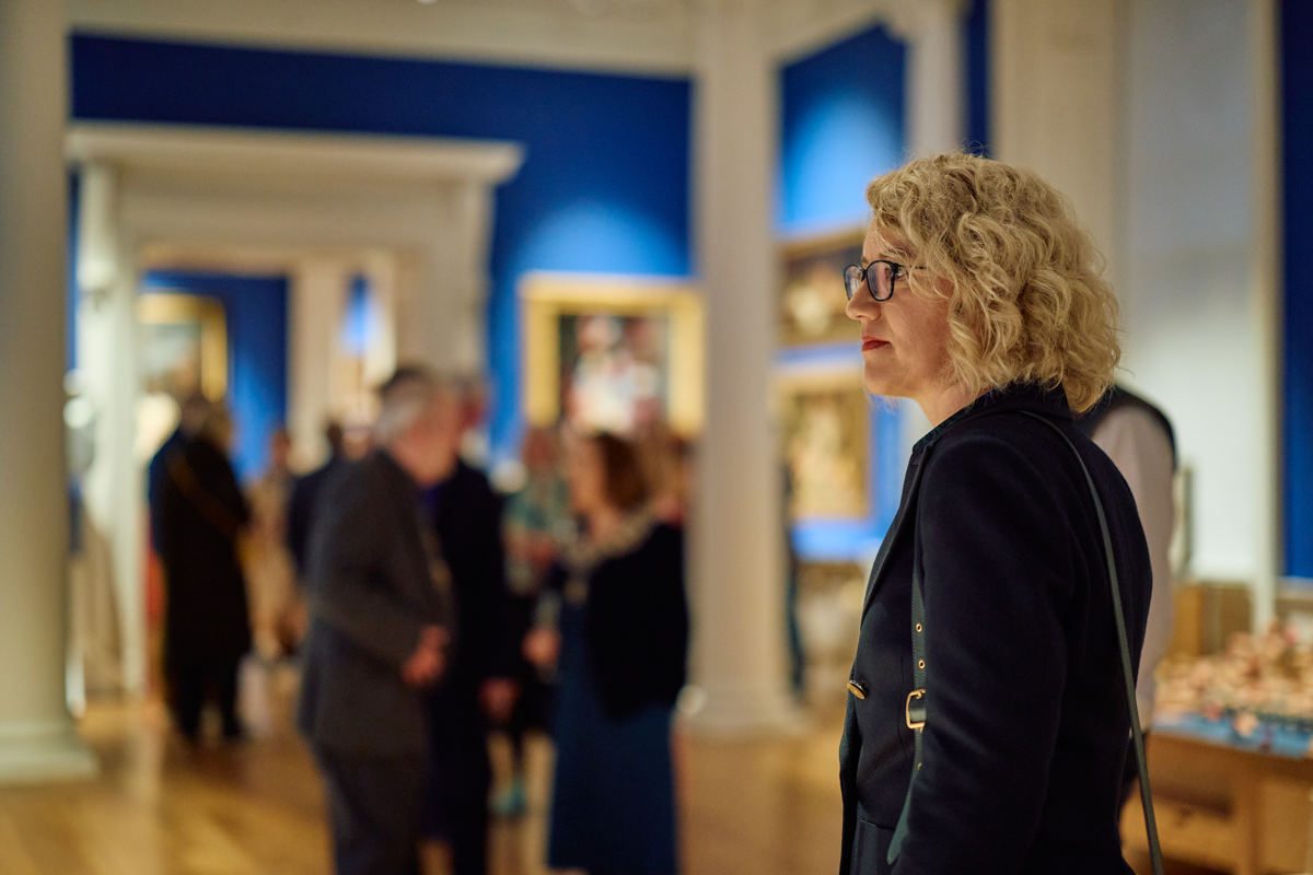 The image shows an art gallery. A woman with curly blonde hair and glasses is standing in the foreground, looking at the artwork on display. She is wearing a black coat or jacket. In the background, there are several other visitors viewing the exhibits, which appear to be framed paintings hanging on blue-coloured walls. The doorways and frames of the gallery space have white trim, creating an artistic and sophisticated environment for displaying and appreciating art.