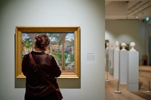 A girl with brown hair tied up with her back to us looks at a large landscape painting in a gallery also lined with sculptures atop plinths.