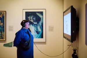 An person wearing headphones and a blue coat is standing in front of a large framed portrait and a display screen in an art exhibition.