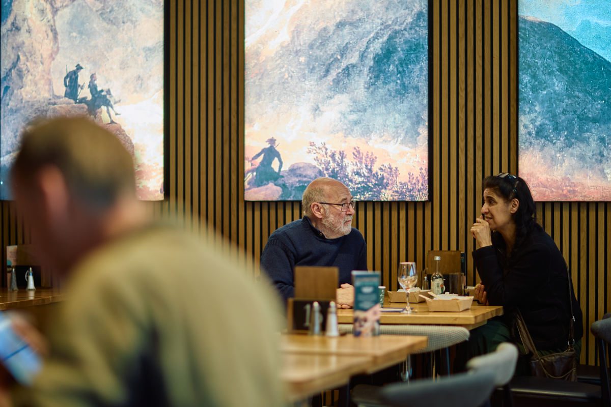 Two peoples speak in a cafe with mugs of hot drink. The interior is wooden panelling and lit-up artworks on the wall.