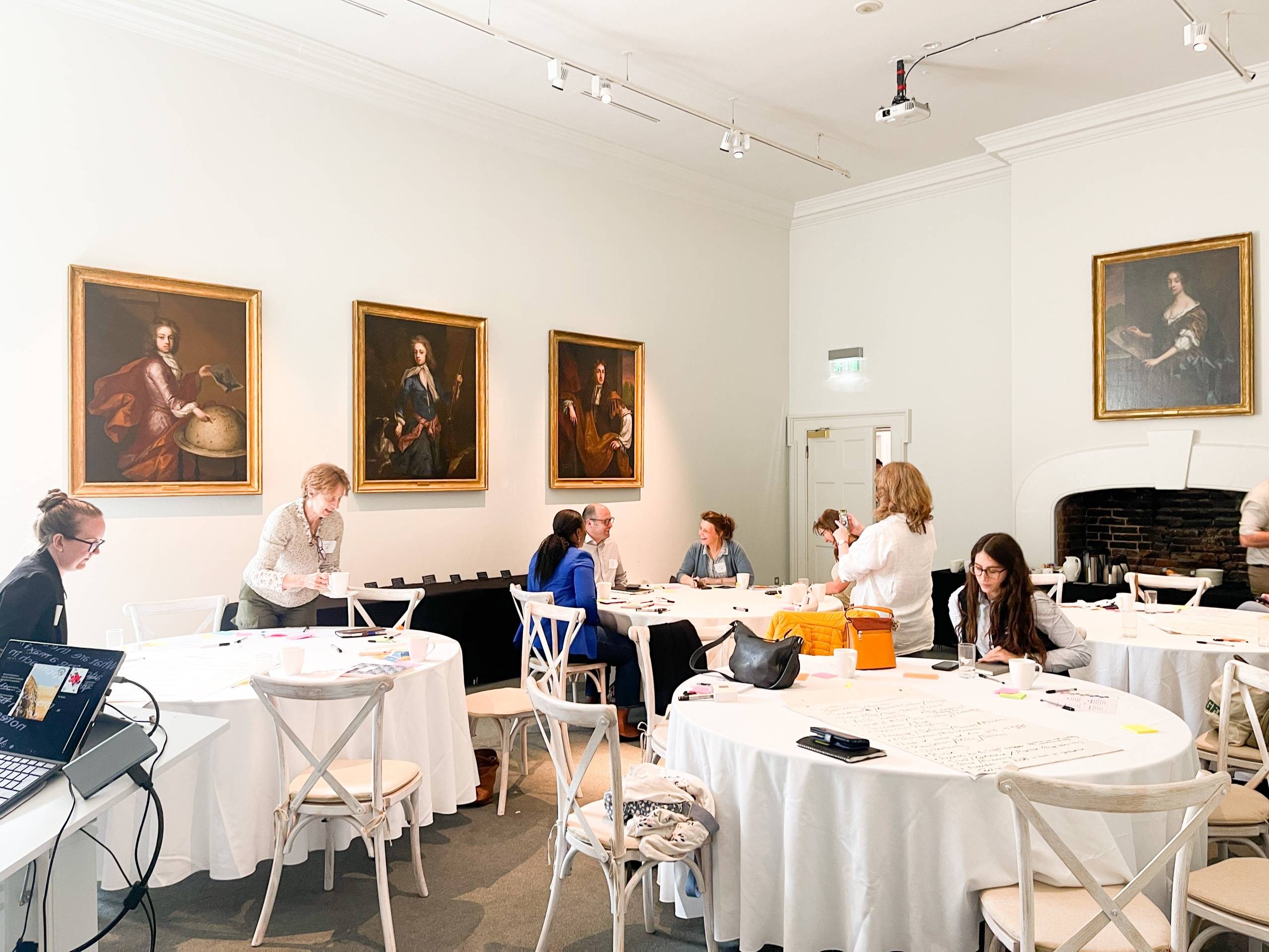 Business people sit around tables talking in a bright white room with a grand fireplace and paintings on the wall.