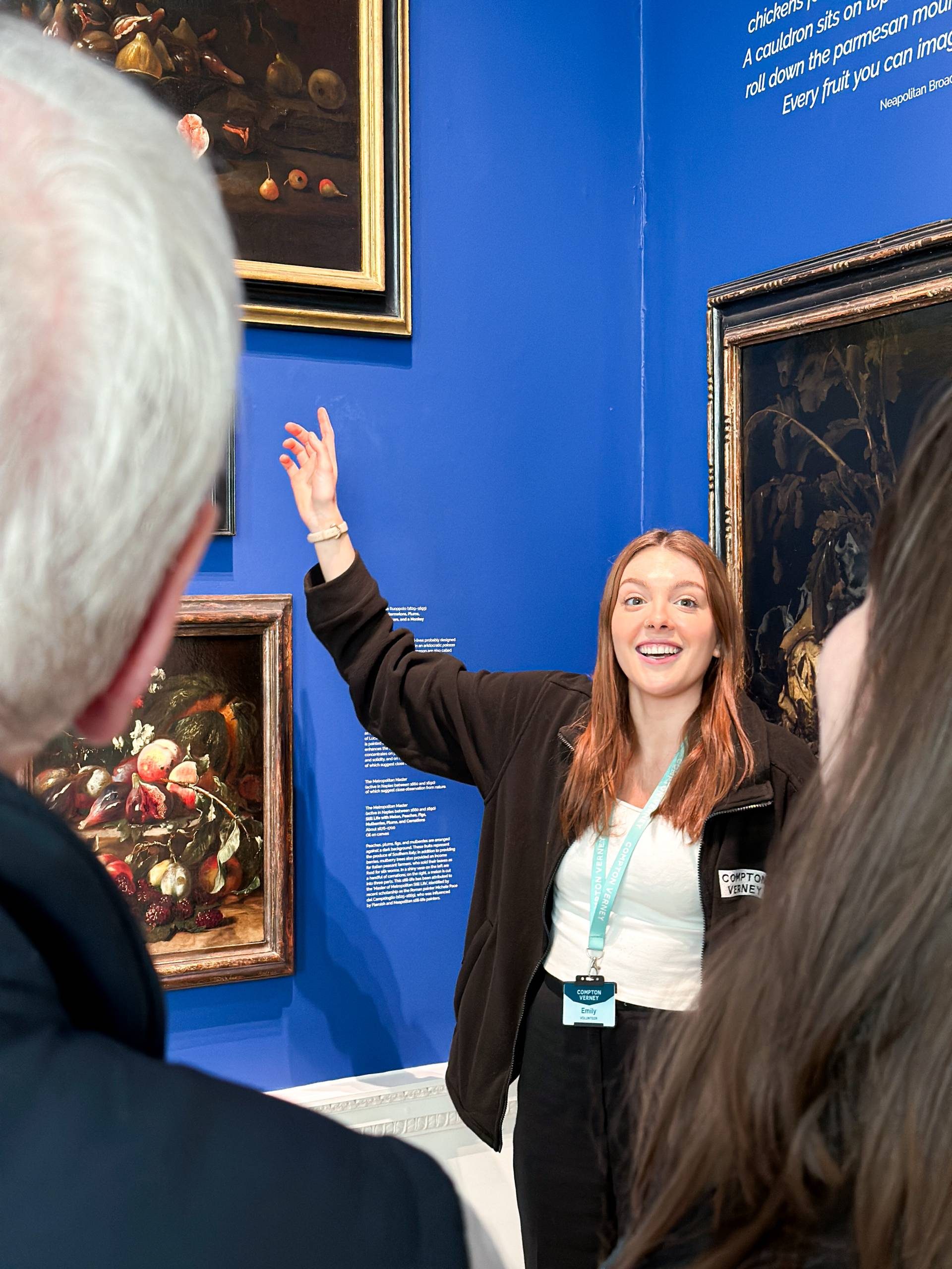 A young female volunteer in Compton Verney uniform points towards a painting on a blue wall infront of two on-lookers.