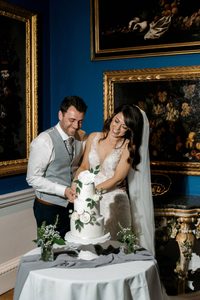 A bridge and groom cut into a three tiered wedding cake in a blue art gallery with floral paintings