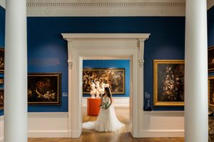 A bride stands holding a bouquet in a bright blue art gallery featuring classical columns, paintings and sculptures with gold frames and red plinths.