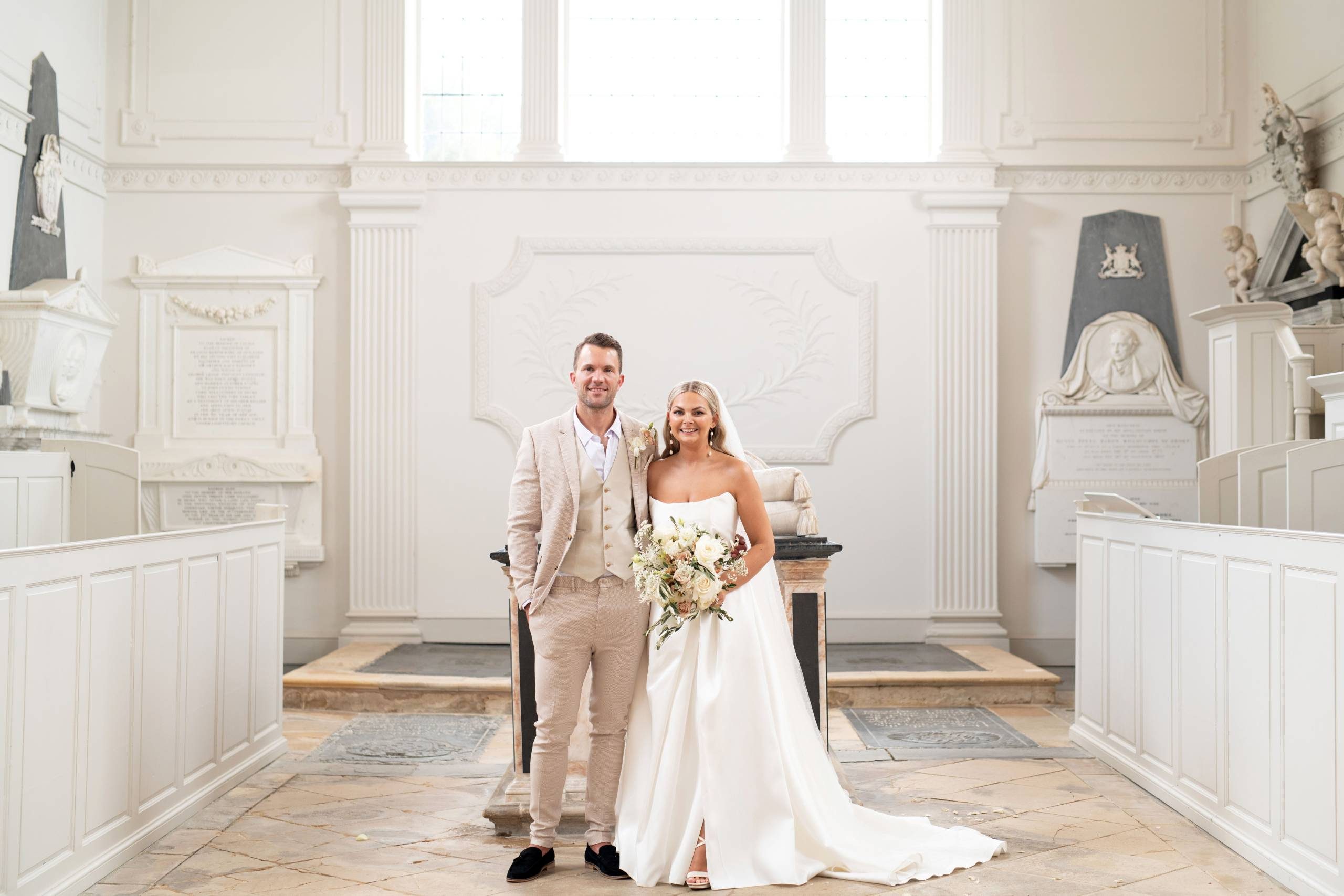A bride and a groom dressed in white and beige stand in an impressive white neoclassical chapel in front of grand light windows