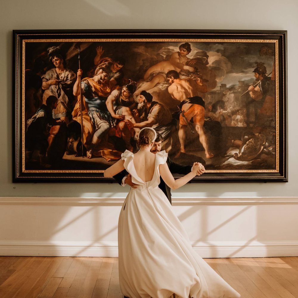 The image depicts a bride in a flowing white dress dancing with a groom in front of a large framed painting in an art gallery. The painting behind her appears to be a dramatic, baroque-style work featuring multiple figures in dynamic poses, possibly depicting a scene from classical mythology or religious narrative. The ornate frame and the woman's graceful movement create an intriguing juxtaposition of the static artwork and the living, dynamic presence in the foreground.