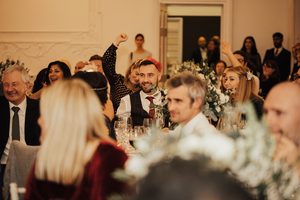 Men and women sit at round tables wearing smart clothes and Christmas cracker hats in a grand white hall