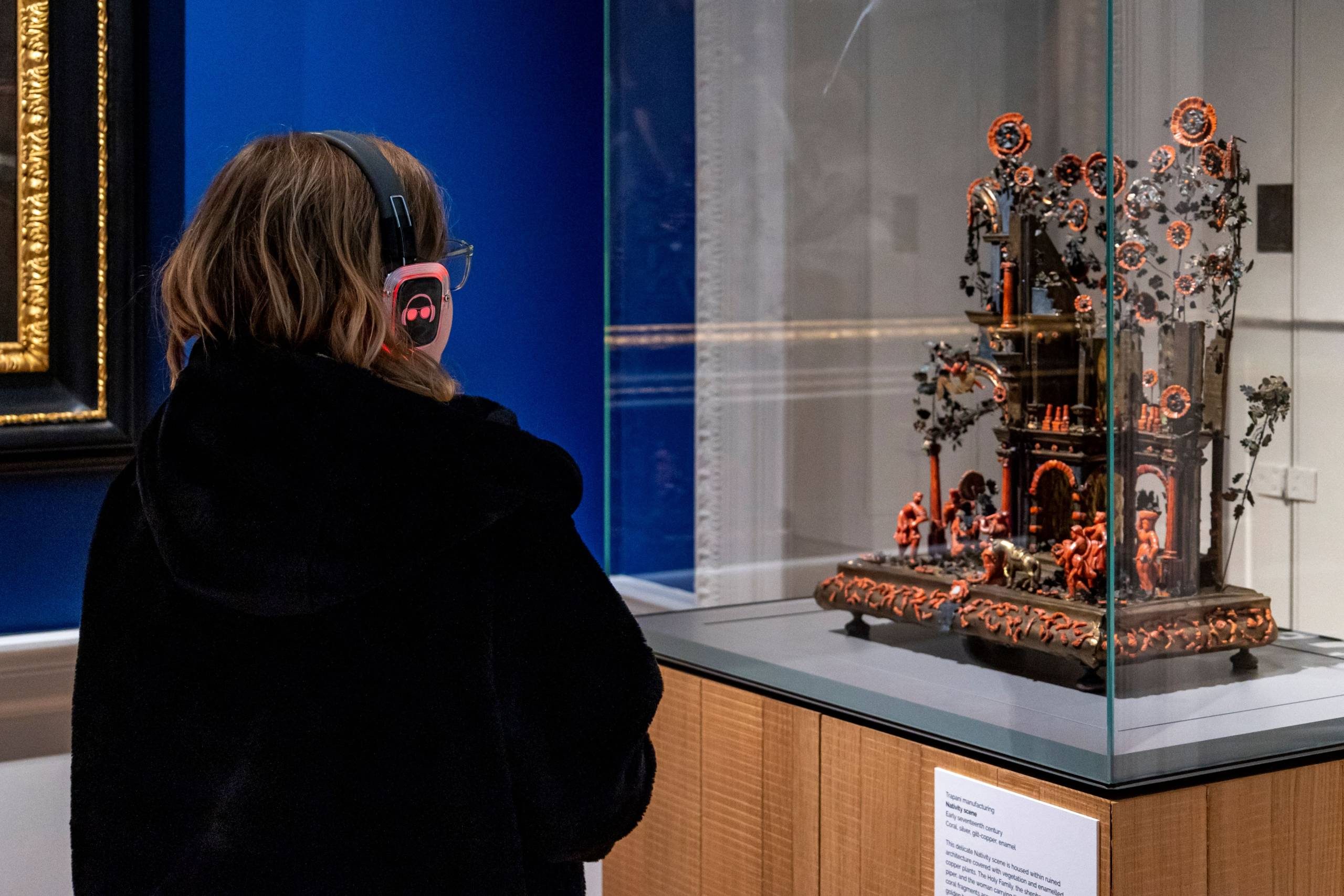 A young girl in a black coat wearing headphones looks at a historical artefact in a glass case in a blue art gallery. The item looks like a religious scene made from bronze and red coral.