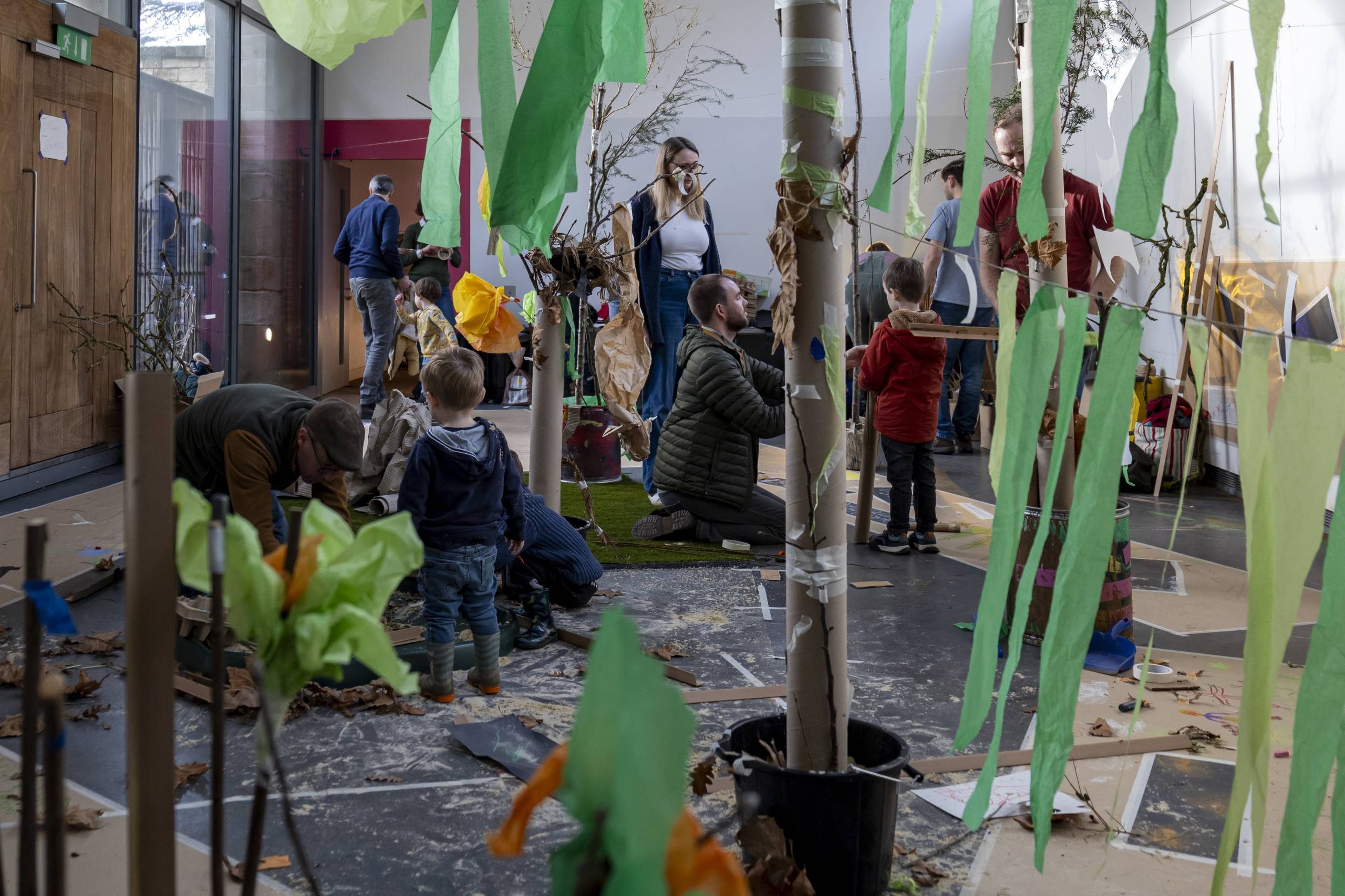 A group of adults and children play in a room filled with paper, leaves, branches and other materials.