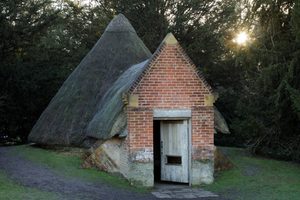 A brick and thatched roof historic ice house with a triangular roof in a woodland setting