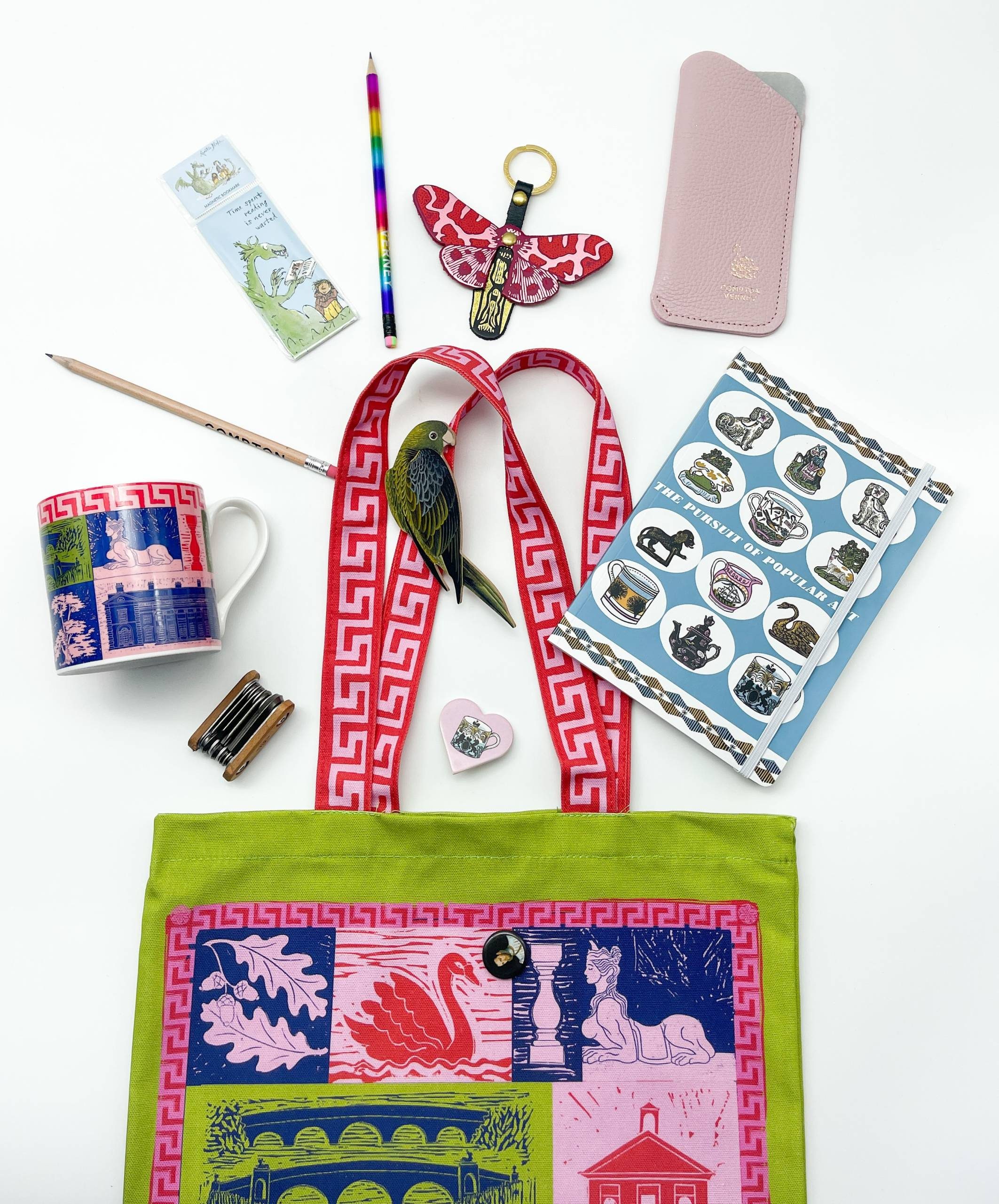 A green, red, and pink tote bag with images of Compton Verney spills open on a white background with Compton Verney gift shop items including a pink glasses case, colourful mug, a bookmark.