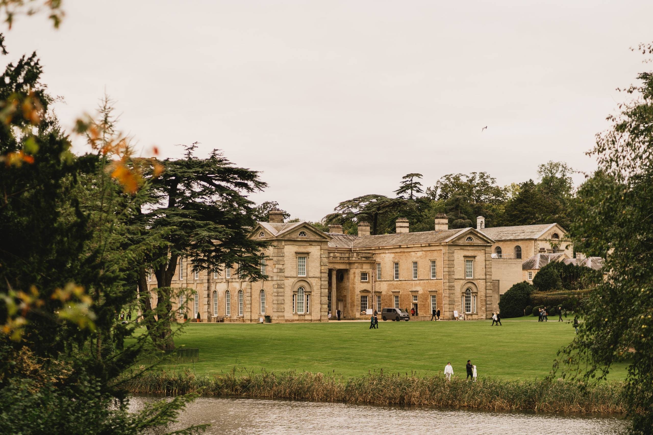 This image shows a historical stone house from across a lake and green meadow with a few people walking and trees framing the image.