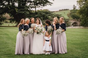 The image shows a bride surrounded by her bridesmaids and a young flower girl in a scenic outdoor setting. The bride is wearing a elegant white gown and holding a bouquet of white flowers, while the bridesmaids are dressed in matching dusty rose-colored dresses and carrying baby's breath bouquets with fur wraps. They are posing together on a grassy field with trees and a rural landscape in the background, creating a romantic and natural backdrop for the bridal party portrait. The group is smiling joyfully, capturing the happiness and camaraderie of the special occasion.