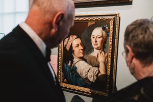 Two people in smart attire look at a portrait of two people in an ornate gold frame in an art gallery.
