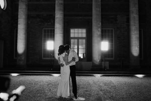 A bride and groom share a loving slow dance in the dark in front of a historical building with large classical columns and bright lights are shining out of the windows hinting at a lively reception going on inside.
