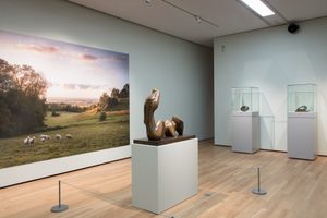 A large photographic mural on one wall showing a scenic landscape with a field, trees, and sheep grazing. In the centre of the room, there is a bronze sculpture displaying an abstract human form on a light pedestal. Along the opposite wall are two tall glass display cases or vitrines, each containing a smaller sculpture.