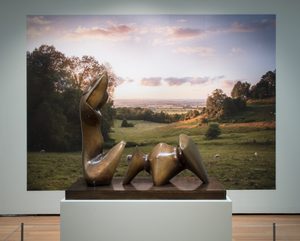 A sculpture of two abstract bronze figures situated on a pedestal in an art gallery setting. The background depicts a peaceful countryside landscape at sunset, with rolling hills, trees, and sheep grazing in a field. The sculpture is the main artistic focus, contrasting with the serene natural scenery behind it. The warm lighting and idyllic vista create an appealing and thoughtful atmosphere for appreciating the artwork.