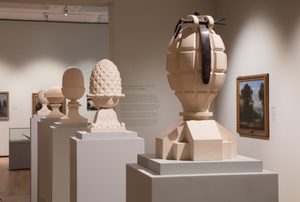 An exhibition gallery displaying various sculptural works. In the foreground, there are several pedestals with ornamental stone sculptures, including spherical and pineapple-shaped forms reminiscent of decorative garden ornaments from the 18th century. The largest sculpture appears to be a stylised depiction of a hand grenade or artillery shell made from pale stone material. The exhibition space also includes framed landscape paintings hanging on the walls.