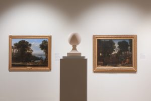 Two framed landscape paintings hung on a wall, with a large stone sculpture in the centre. The sculpture is an abstract orb form on a rectangular pedestal. The paintings depict scenic outdoor views with trees and bodies of water.
