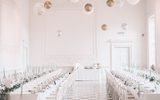 This image shows a grand classical hall filled with long tables, chairs, green florals ready for a wedding reception. Large gold and white balloons float up to the stuccoed ceiling.