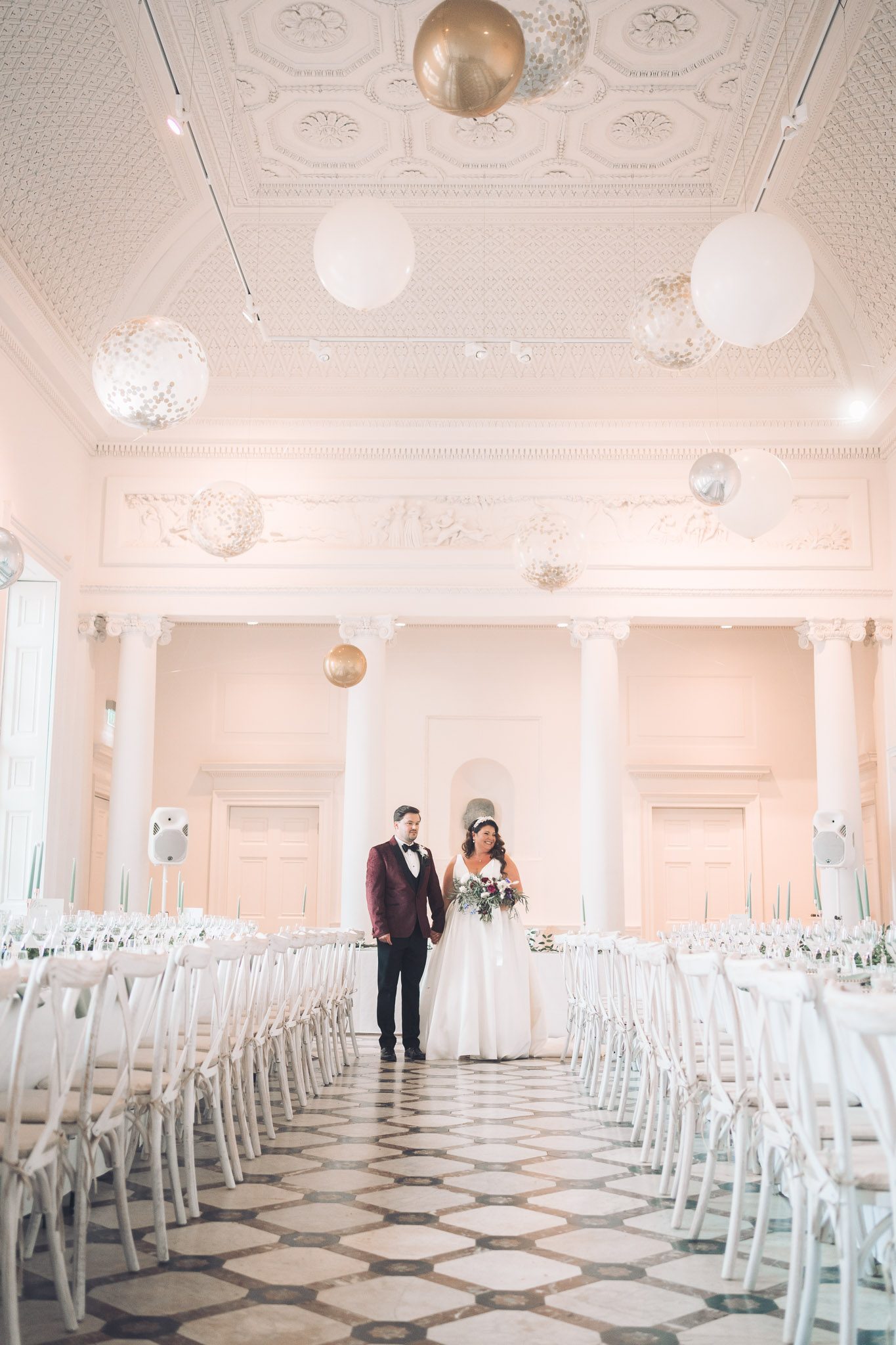 A bride and groom in white and burgundy stand in a grand white hall filled with large white and gold balloons, tables and chairs, and flowers.