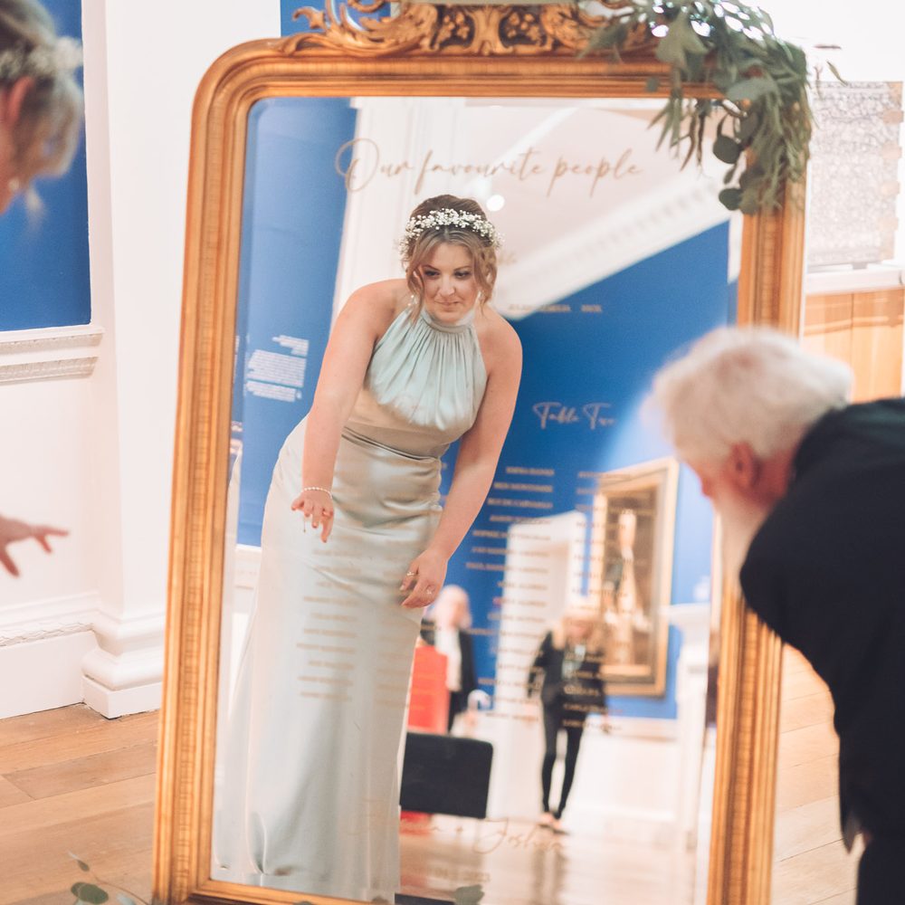 A bridesmaid in a green dress points at a seating chart written in gold in a mirror that reflects the blue art gallery around