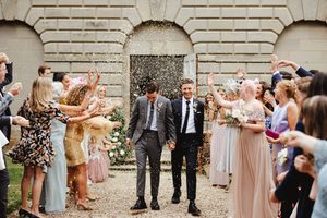 Two grooms walks through confetti after leaving a historic chapel, smiling as family and friends in formal dress surround them.