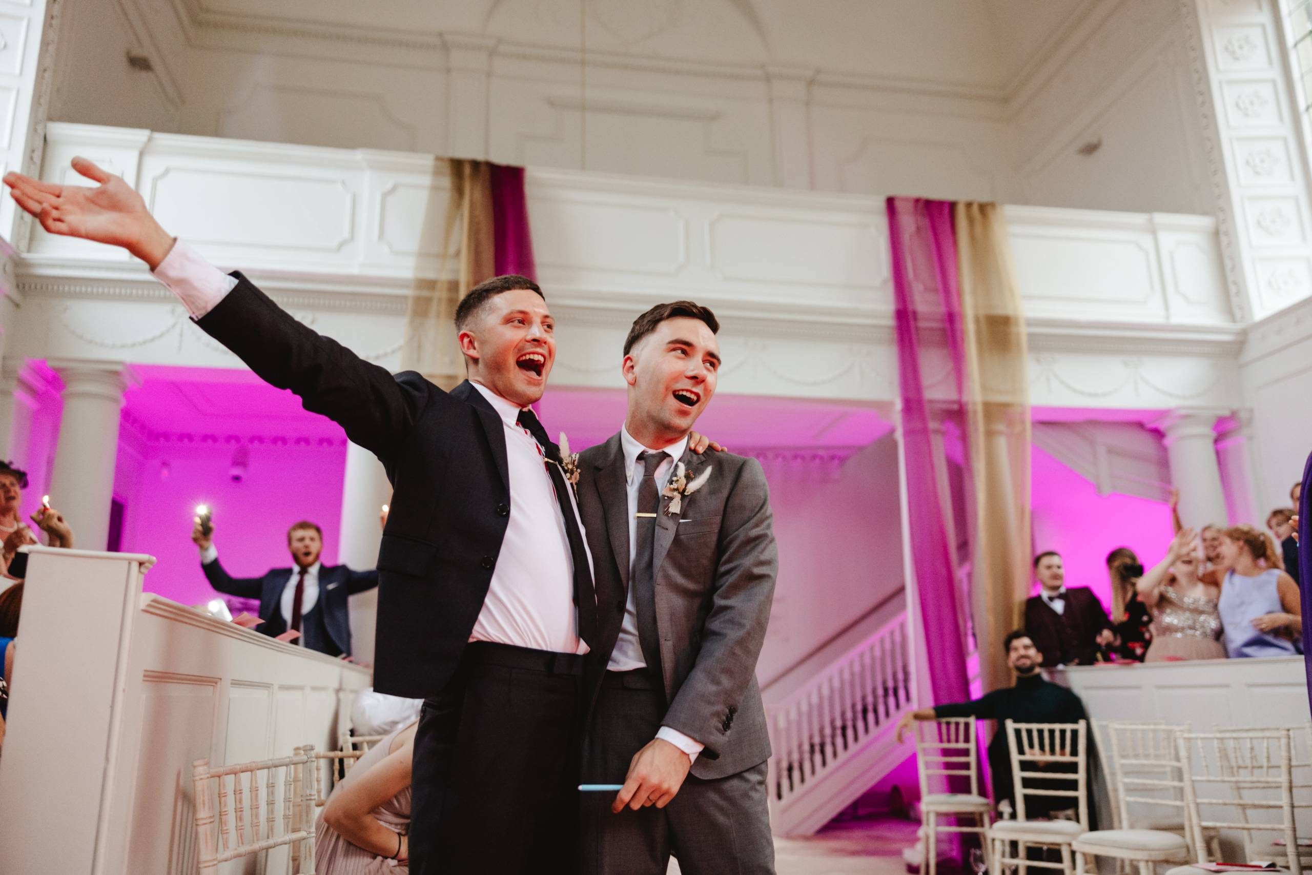 Two grooms in black suits celebrate in a white neoclassical chapel draped with colourful fabric