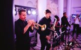 A live band with trombones and saxophones performs in a classical hall with purple lights.