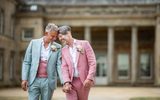 The image shows a loving and joyful same-sex couple holding hands and embracing after their wedding ceremony. They are wearing complementary pastel-colored suits accented with boutonniere flowers, suggesting they exchanged vows in a formal setting. Their beaming smiles and physical closeness radiates their happiness and commitment to one another. The background features a classical building with columns and stone features