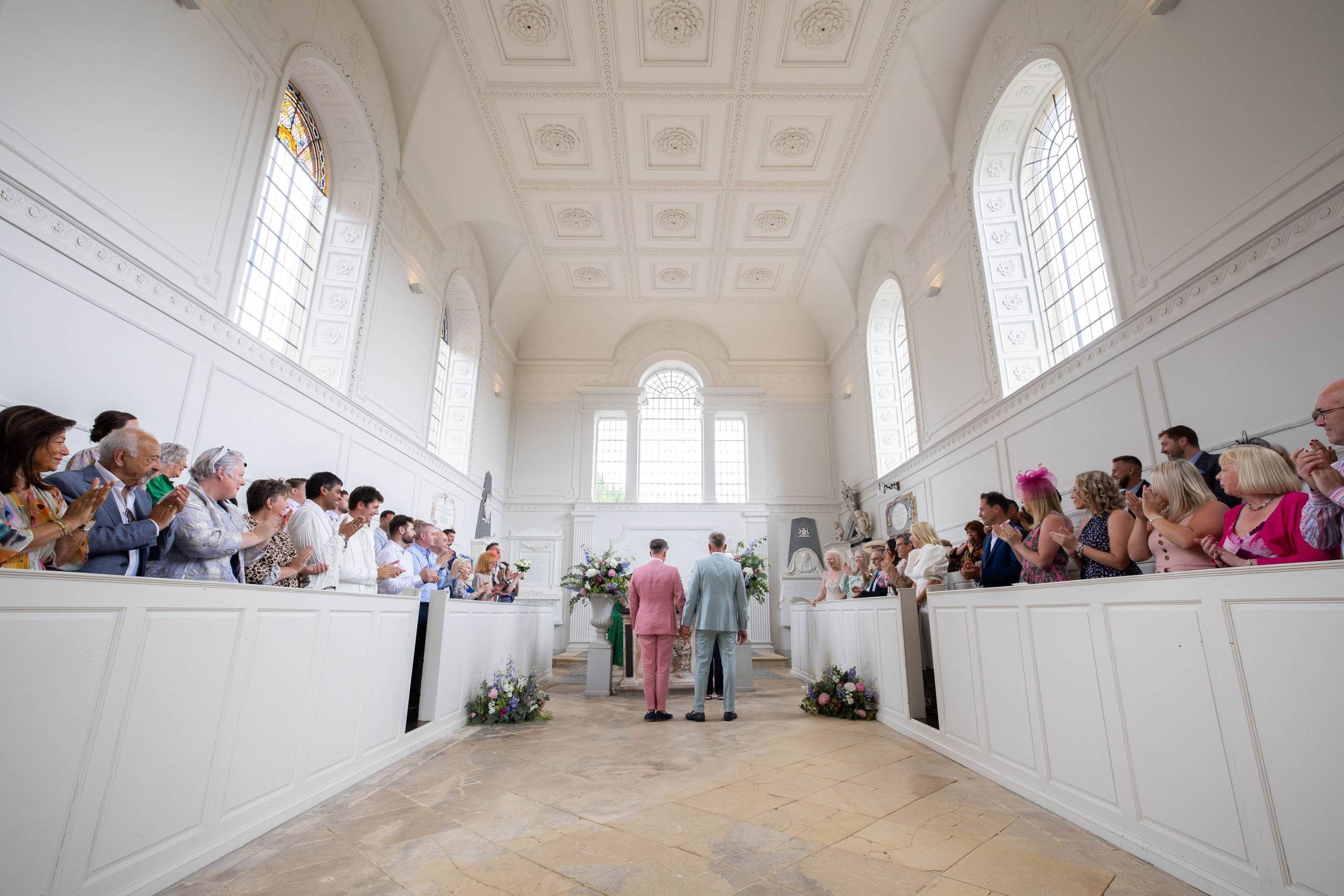 A groom in a blue suit and a groom in a pink suit stand at an altar in a white neo-classical chapel