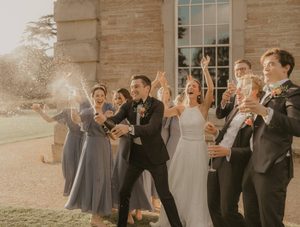 A bridal party made up of the bridge, groom, bridesmaids and groomsmen pop champagne in front of a historical stone building as the sun sets behind them.