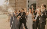 A bridal party made up of the bridge, groom, bridesmaids and groomsmen pop champagne in front of a historical stone building as the sun sets behind them.